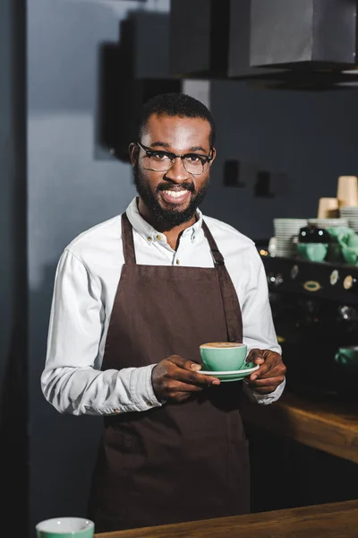 Beau barista afro-américain aux lunettes tenant une tasse de cappuccino et souriant à la caméra — Photo de stock