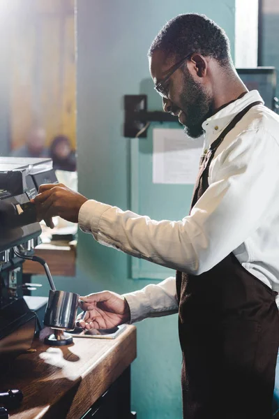 Vista lateral de la joven barista afroamericana feliz en anteojos haciendo café en la máquina de café - foto de stock