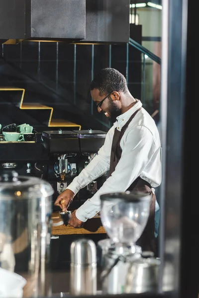 Side view of smiling young african american barista in eyeglasses making coffee at coffee machine — Stock Photo