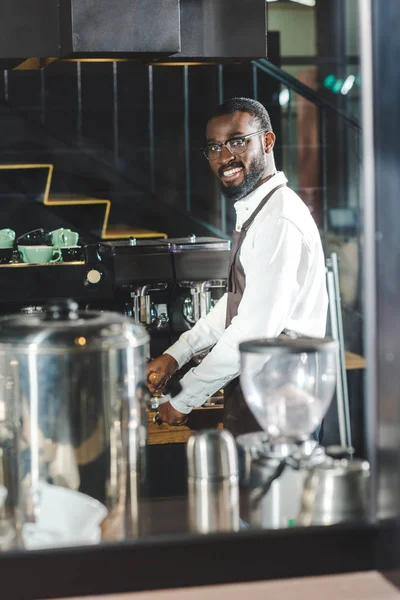 Красивий молодий афроамериканець бариста в окулярів, посміхаючись на камеру під час приготування кави на каву-машиною — стокове фото
