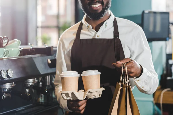 Colpo ritagliato di sorridente barista africano americano che tiene sacchetti di carta e tazze di caffè usa e getta in caffè — Foto stock