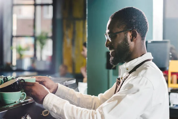 Vista lateral de sonriente joven africano americano barista sosteniendo tazas y trabajando con la máquina de café en la cafetería - foto de stock