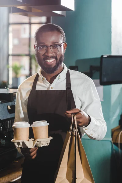 Allegro giovane barista afroamericano in possesso di tazze di carta e borse in caffetteria — Foto stock