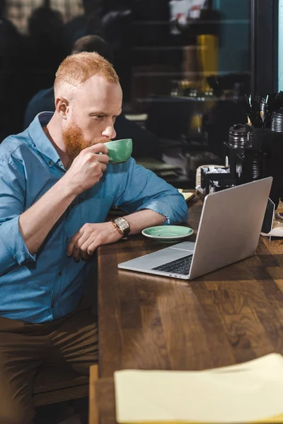 Schöner bärtiger junger Mann benutzt Laptop und trinkt Kaffee im Café — Stockfoto