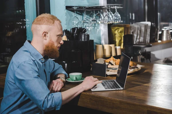 Вид сбоку красивого бородатого молодого человека с ноутбуком с экраном в кофейне — стоковое фото