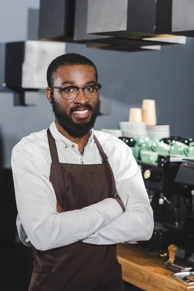 Confiant barista afro-américain dans des lunettes debout avec les bras croisés et souriant à la caméra dans un café — Photo de stock