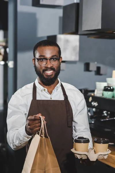 Junge afrikanisch-amerikanische Barista hält Einweg-Kaffeetassen und Papiertüten in der Hand und lächelt in die Kamera im Café — Stockfoto