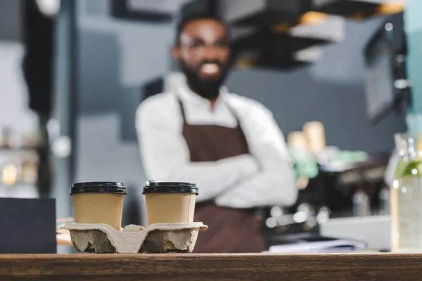 Vista ravvicinata di due tazze di carta con caffè e barista afroamericano sorridente in piedi con le braccia incrociate dietro — Foto stock