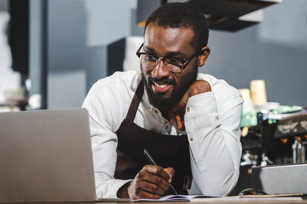 Bonito jovem africano americano barista tomar notas e usando laptop no café — Fotografia de Stock