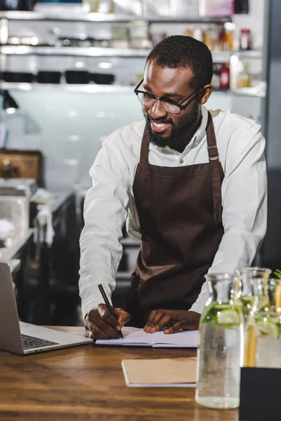 Feliz joven afroamericano barista tomando notas y el uso de la computadora portátil en la cafetería - foto de stock