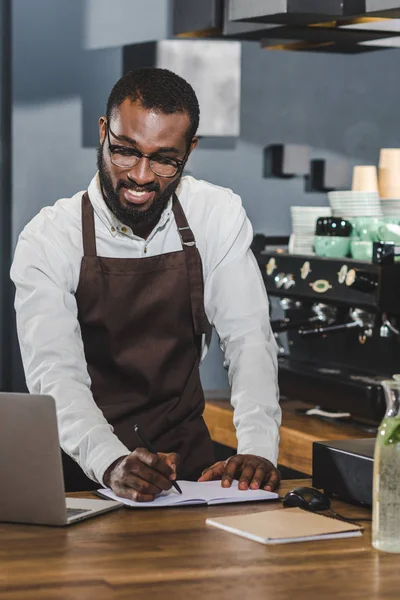 Sonriente joven afroamericano barista tomando notas y el uso de la computadora portátil en la cafetería - foto de stock