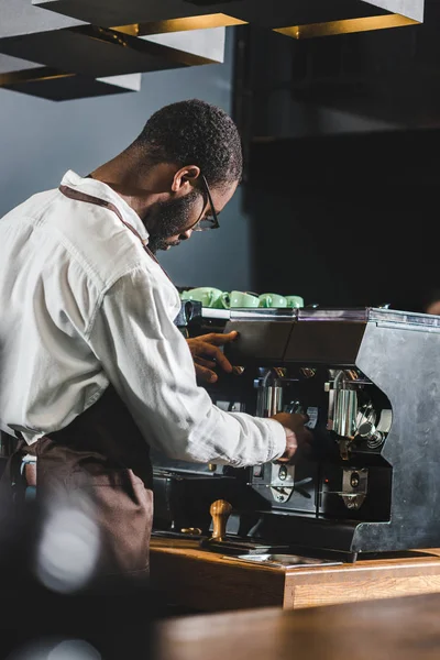 Joven barista afroamericano en anteojos y delantal trabajando con máquina de café - foto de stock