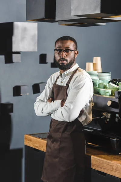 Confiant barista afro-américain dans des lunettes debout avec les bras croisés et regardant la caméra dans le café — Photo de stock
