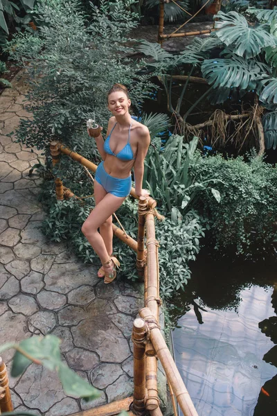 Blick von oben auf eine schlanke junge Frau im Bikini, die mit Kokoscocktail im tropischen Garten posiert — Stockfoto