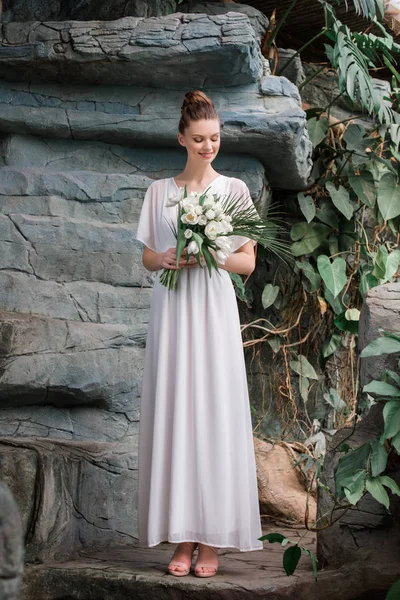 Novia sonriente posando en vestido blanco con ramo de bodas en jardín tropical - foto de stock