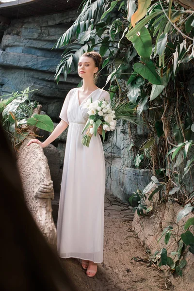 Hermosa novia posando en vestido blanco con ramo de bodas en jardín tropical - foto de stock