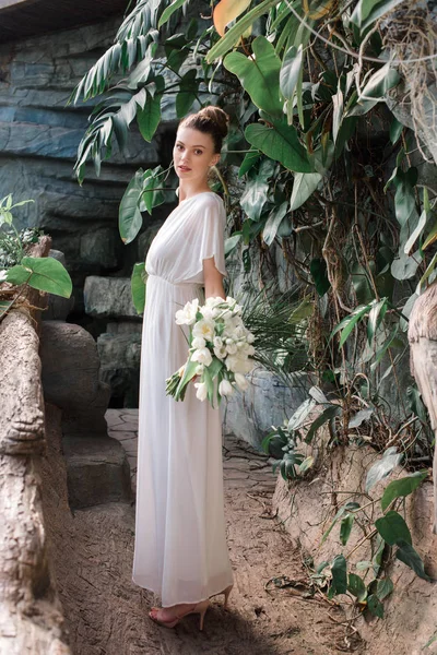 Bonita novia posando en vestido blanco con ramo de bodas en jardín tropical - foto de stock