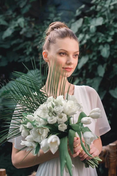 Sposa felice posa in abito bianco con bouquet da sposa nel giardino tropicale — Foto stock