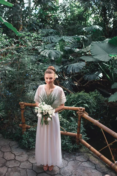 Joven novia posando en vestido blanco con ramo de bodas en jardín tropical - foto de stock