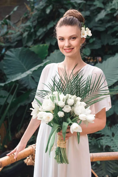 Lächelnde Braut posiert im weißen Kleid mit Brautstrauß im tropischen Garten — Stockfoto