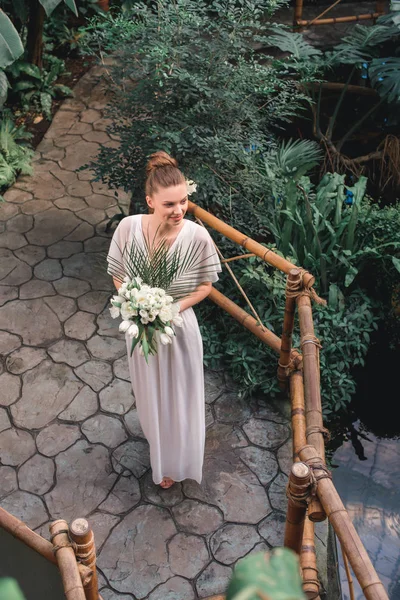 Vue aérienne de jolie mariée posant en robe blanche avec bouquet de mariage dans le jardin tropical — Photo de stock