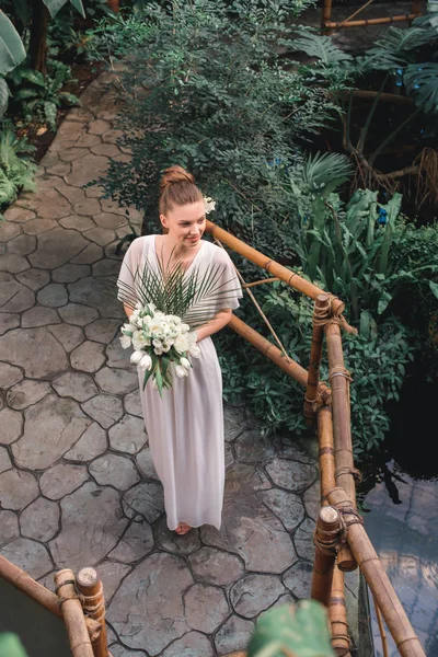 Overhead view of pretty bride posing in white dress with wedding bouquet in tropical garden — Stock Photo