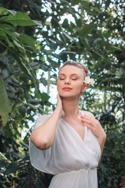 Hübsche Frau im weißen Kleid mit geschlossenen Augen und Blume im Haar posiert im tropischen Garten — Stockfoto