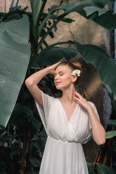 Нежная женщина в белом платье с цветком в волосах позирует в тропическом саду — стоковое фото