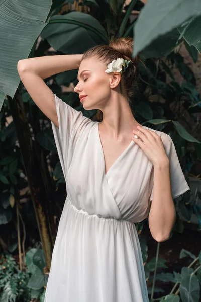 Atractiva novia tierna en vestido blanco con flor en el pelo posando en el jardín tropical - foto de stock