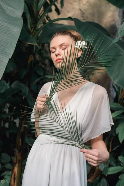 Tierna chica en vestido de verano blanco posando con hojas de palma tropical - foto de stock