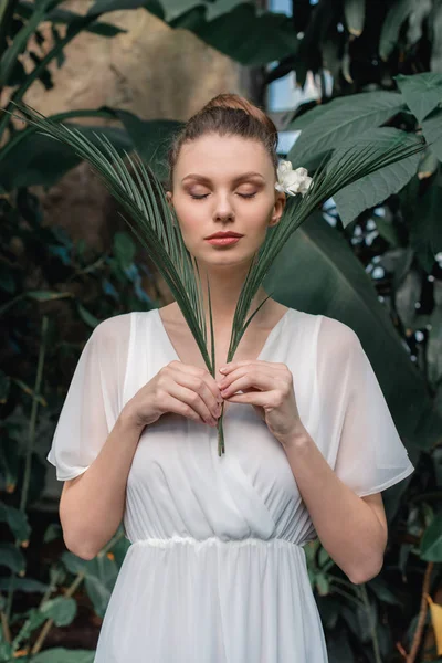 Attraktives Mädchen im weißen Sommerkleid posiert mit geschlossenen Augen und tropischen Palmenblättern — Stockfoto