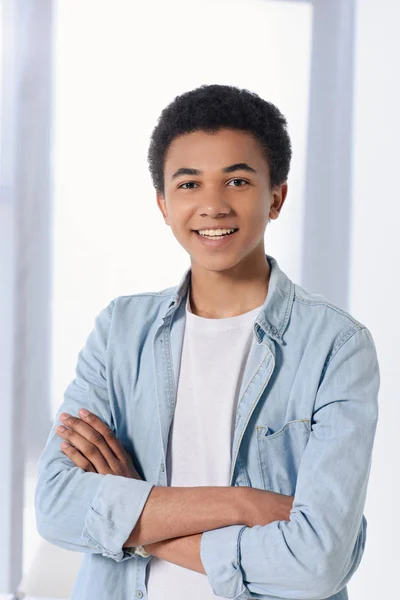 Sonriente afroamericano adolescente de pie con los brazos cruzados y mirando a la cámara en casa - foto de stock