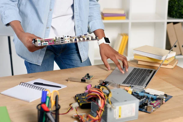Cropped image of african american teenager holding computer motherboard and using laptop at home — Stock Photo