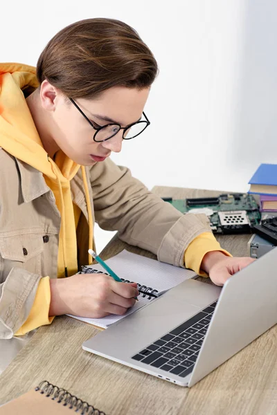 Teen boy using laptop and writing something to notebook at home — Stock Photo