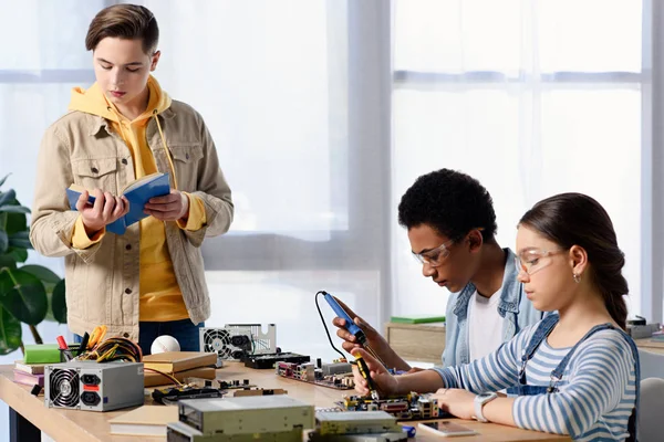 Multicultural teenagers soldering computer circuit with soldering iron and friend reading book — Stock Photo