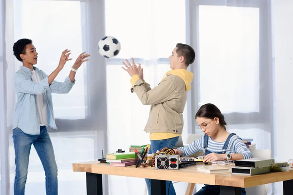 Multicultural adolescente chicos jugando con fútbol pelota mientras femenino adolescente fijación circuito en casa - foto de stock