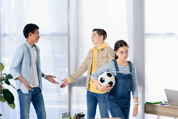 Multicultural teenagers showing shrug gestures while friends walking away with football ball at home — Stock Photo