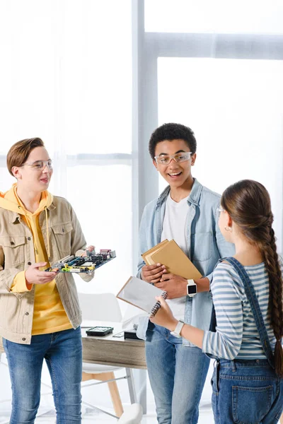 Adolescentes multiétnicos sosteniendo la placa madre de la computadora y los libros en casa - foto de stock