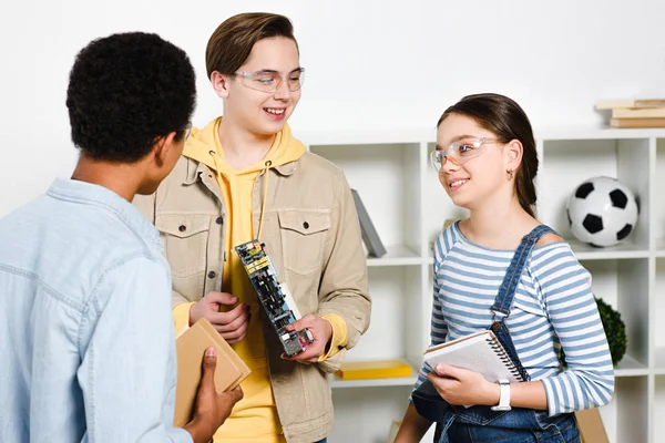 Multicultural teenagers talking and holding computer motherboard and books at home — Stock Photo