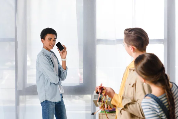 Chico afroamericano hablando por teléfono inteligente y mirando a los amigos en casa - foto de stock