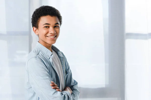 Улыбающийся африканский американский подросток, стоящий со скрещенными руками и смотрящий в камеру дома — стоковое фото