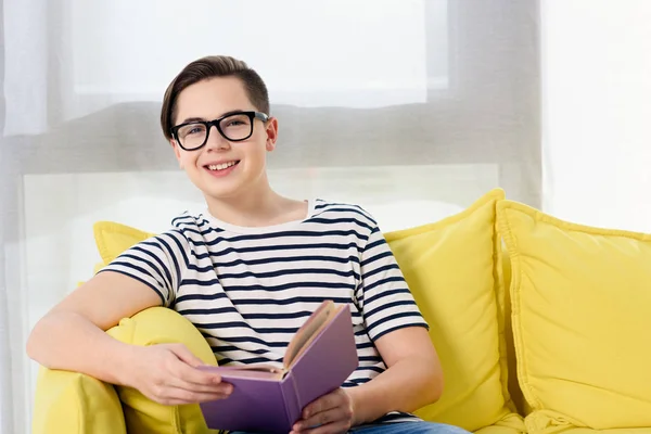 Sorrindo adolescente menino sentado com livro violeta no sofá amarelo em casa — Fotografia de Stock
