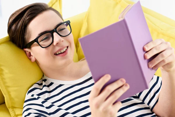 Sonriente adolescente chico leyendo violeta libro en casa - foto de stock