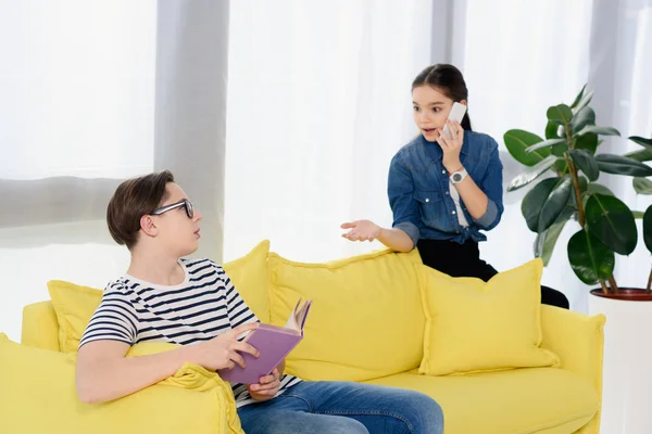 Teenager talking by smartphone and looking at boy sitting on sofa with book at home — Stock Photo