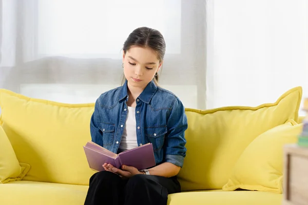 Adorable preteen child sitting on yellow sofa and reading book at home — Stock Photo