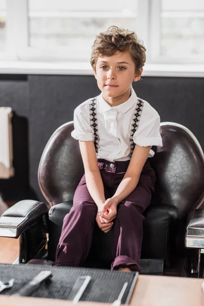 Niño rizado sentado en la silla de barbero en la barbería - foto de stock