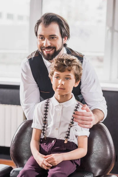 Adorable little kid sitting on chair at barbershop while happy barber standing behind him — Stock Photo