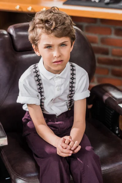 Високий кут зору стильна маленька дитина сидить у перукарському кріслі в перукарні і дивиться на камеру — стокове фото