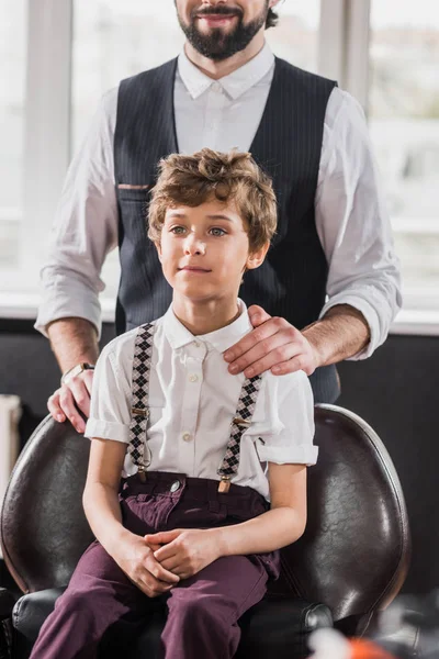 Adorable petit enfant assis sur la chaise au salon de coiffure tandis que le coiffeur debout derrière lui — Photo de stock