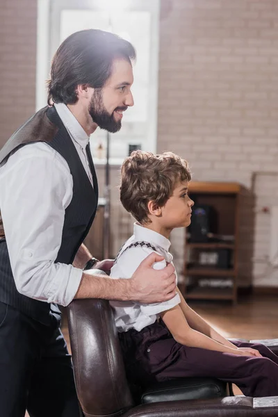 Seitenansicht des hübschen Friseurs mit Blick auf das Spiegelbild eines kleinen Kindes, das im Stuhl beim Friseur sitzt — Stockfoto